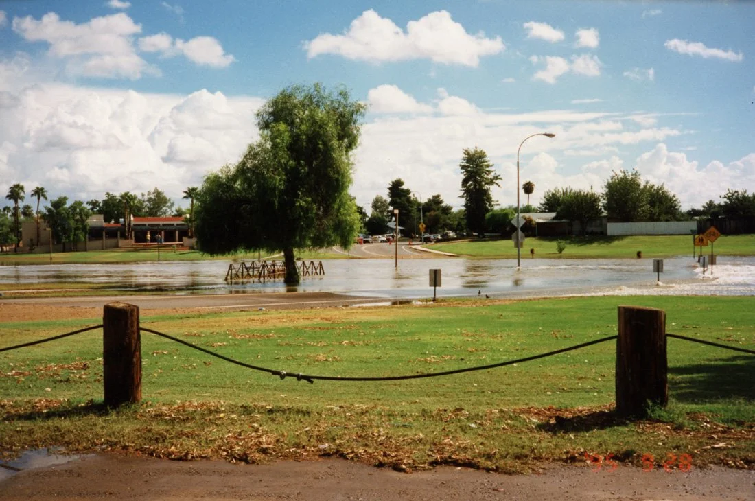 Floodwaters submerge a park area with a tree, street signs, and a fence, while water covers the road and extends into the background.