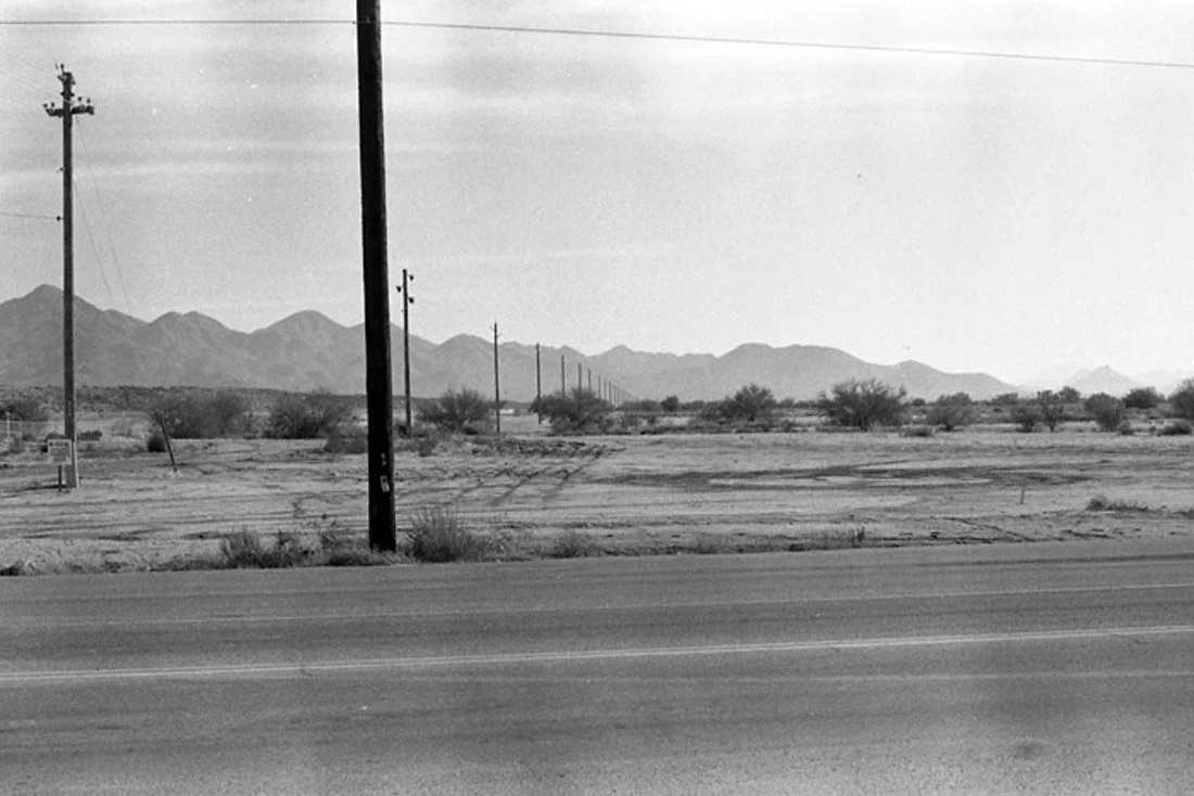 Black and white photo of a desert landscape with mountains in the distance, power lines and utility poles running parallel to the road in the foreground.