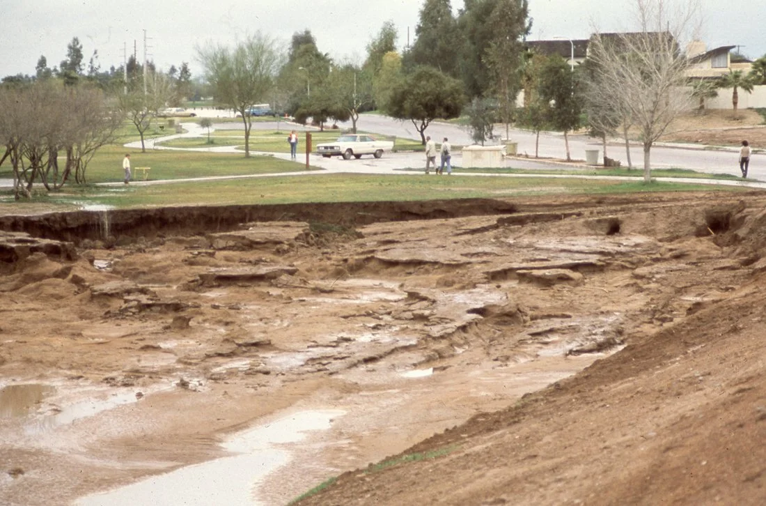 A large sinkhole in a park with eroded soil and muddy water, surrounded by trees and a paved walkway. People are walking in the background.