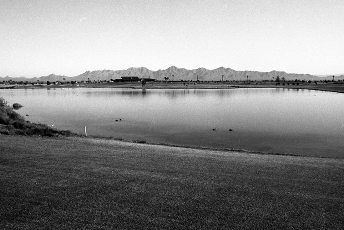 A black and white photo of a lake with a mountainous landscape in the background, and a flat grassy area in the foreground.