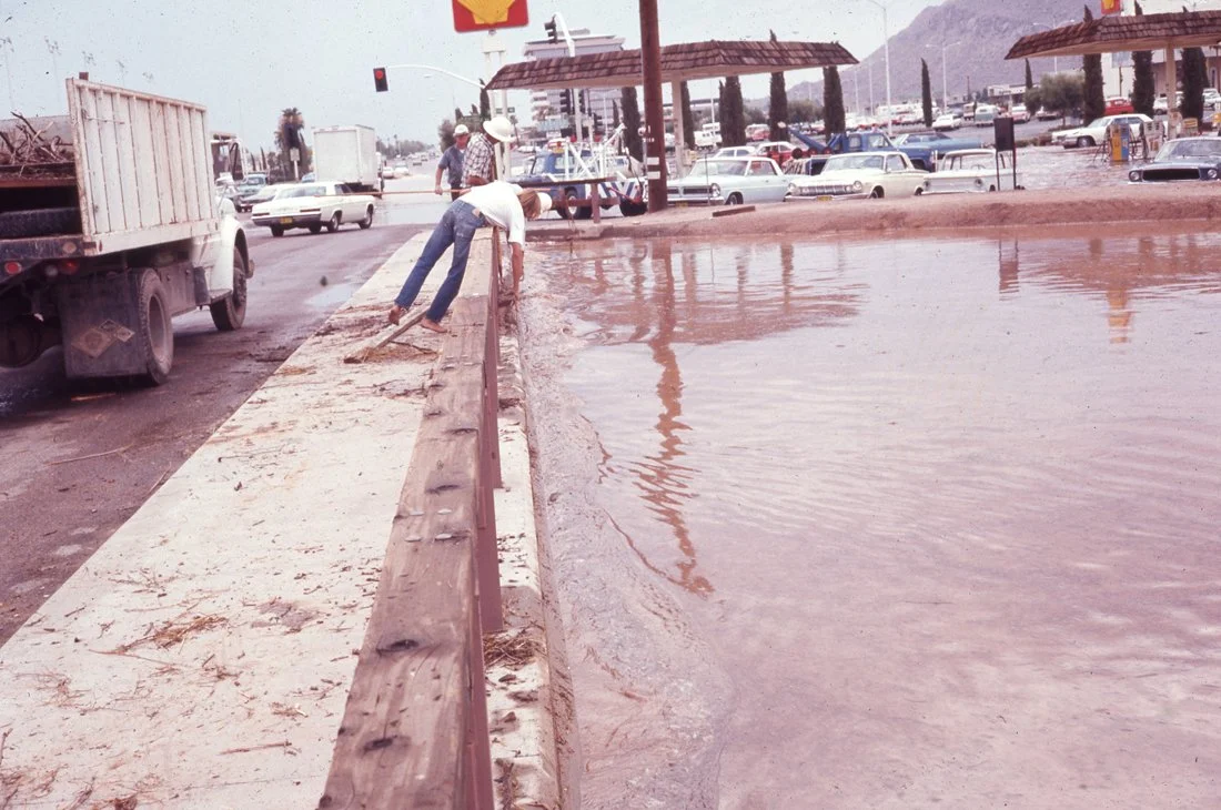 A man cleaning a flooded roadside with a broom next to a guardrail, near a gas station with cars and trucks in the background.