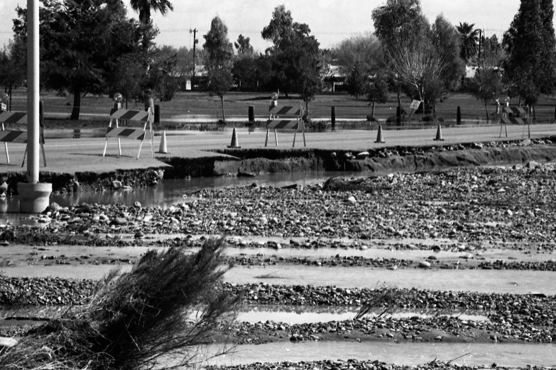 A black and white photo of a street with construction barriers, warning cones, damaged pavement, and a small water stream, with trees and houses in the background.