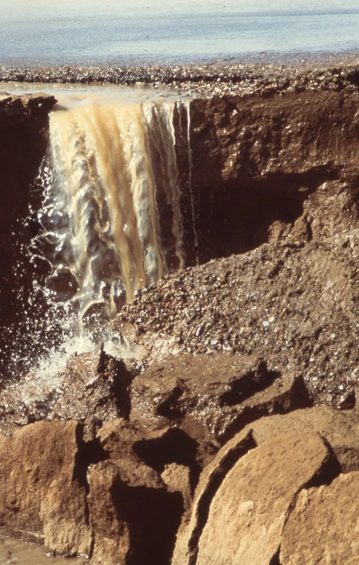 A small waterfall flowing over rocks into a body of water, with rocks in the foreground and water in the background.