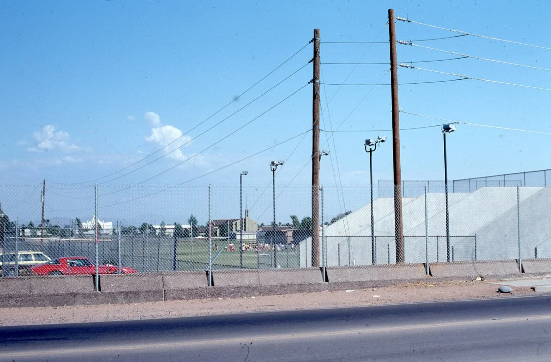 Fenced sports field with floodlights, utility poles, parked cars, and sidewalk under a partly cloudy sky.