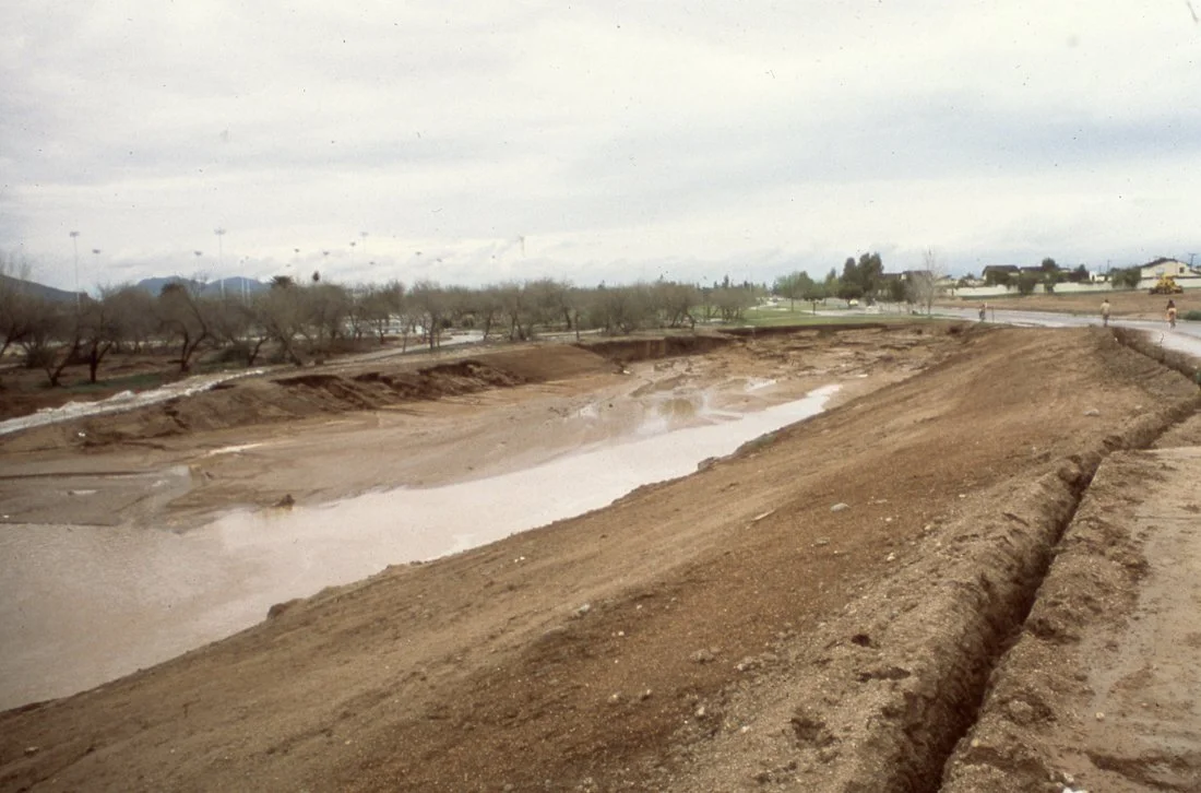 A construction site with dirt mounds and a partially excavated area with muddy water, overcast sky, and some trees and buildings in the background.