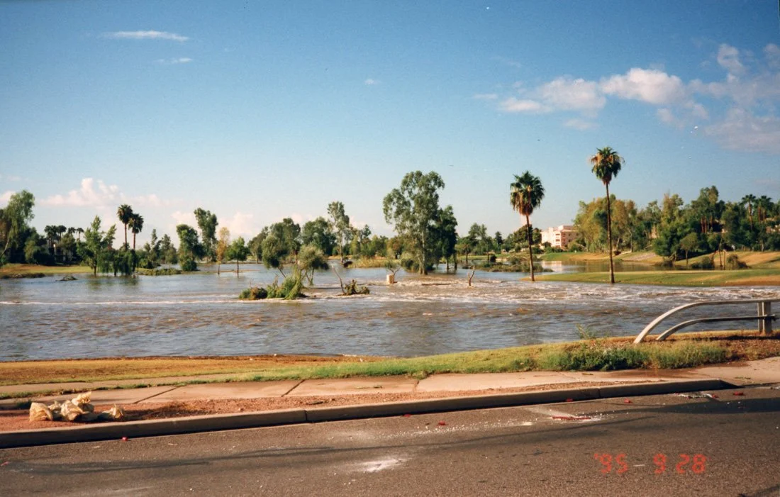 Flooded area with water overflowing onto the street, trees partially submerged, and a blue sky with clouds.