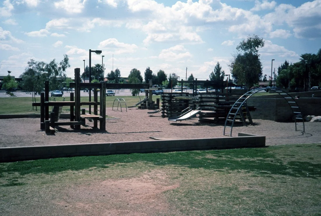 Empty playground with playground equipment, slides, climbing structures, and a grassy area in the foreground under cloudy sky.