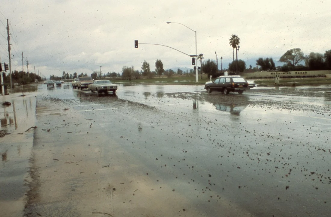 Cars driving through a flooded intersection during rainy weather with puddles and wet roads, some traffic lights visible, palm trees in the background.