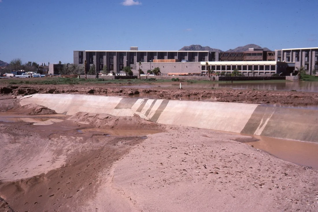 Construction site with a partially built dam or retaining wall in front of a modern building with mountains in the background.