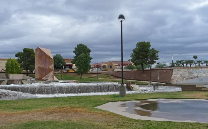 A park with a small waterfall feature, a pond, trees, a lamppost, and buildings in the background on a cloudy day.
