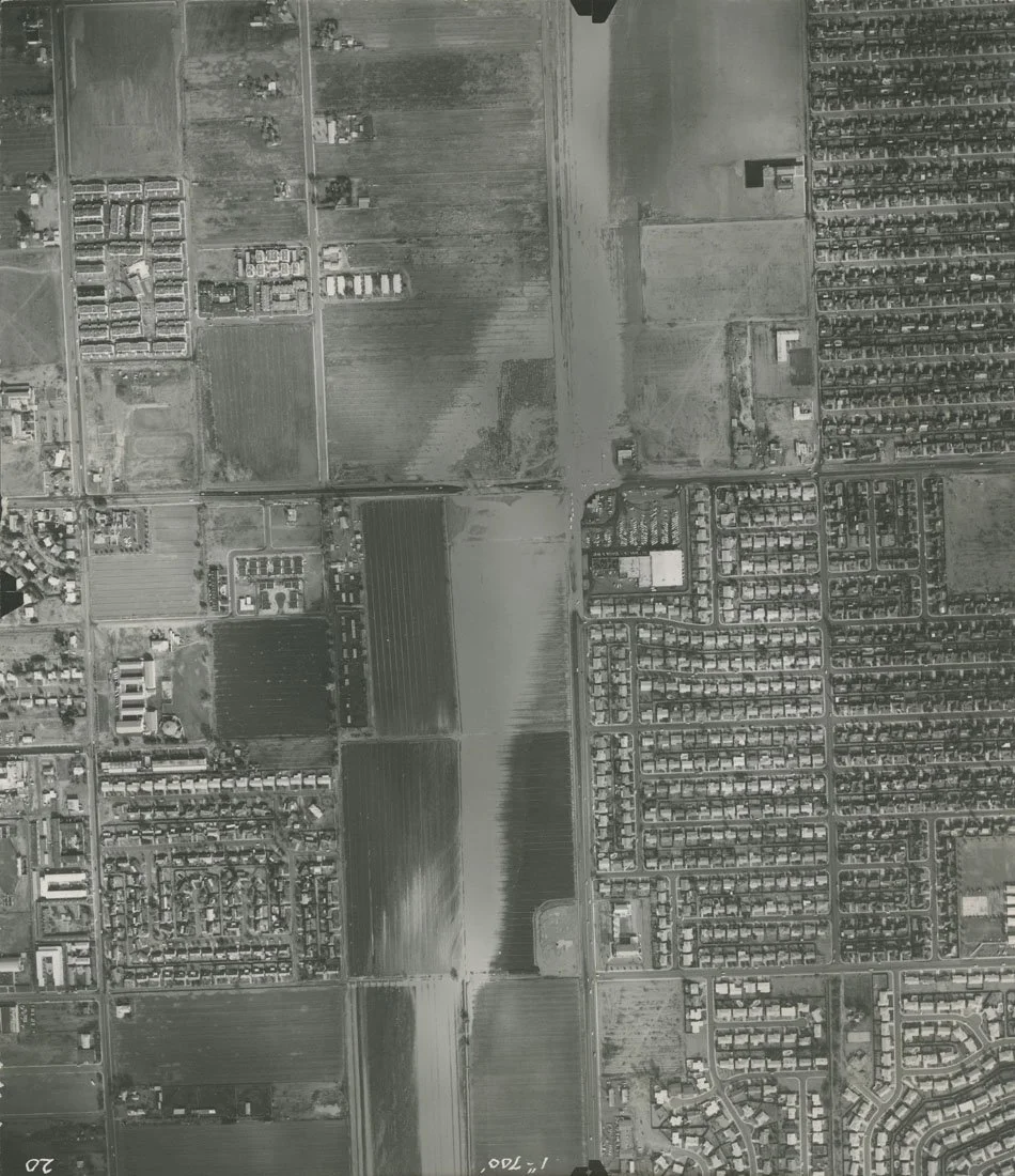Aerial black-and-white satellite image showing residential neighborhoods, farmland, and open fields, with grid-like street patterns and some rural structures.