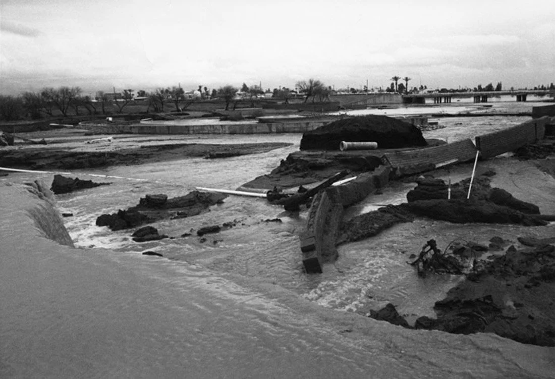 Black and white photo of flood damage with displaced soil, damaged infrastructure, and debris in a flooded area.