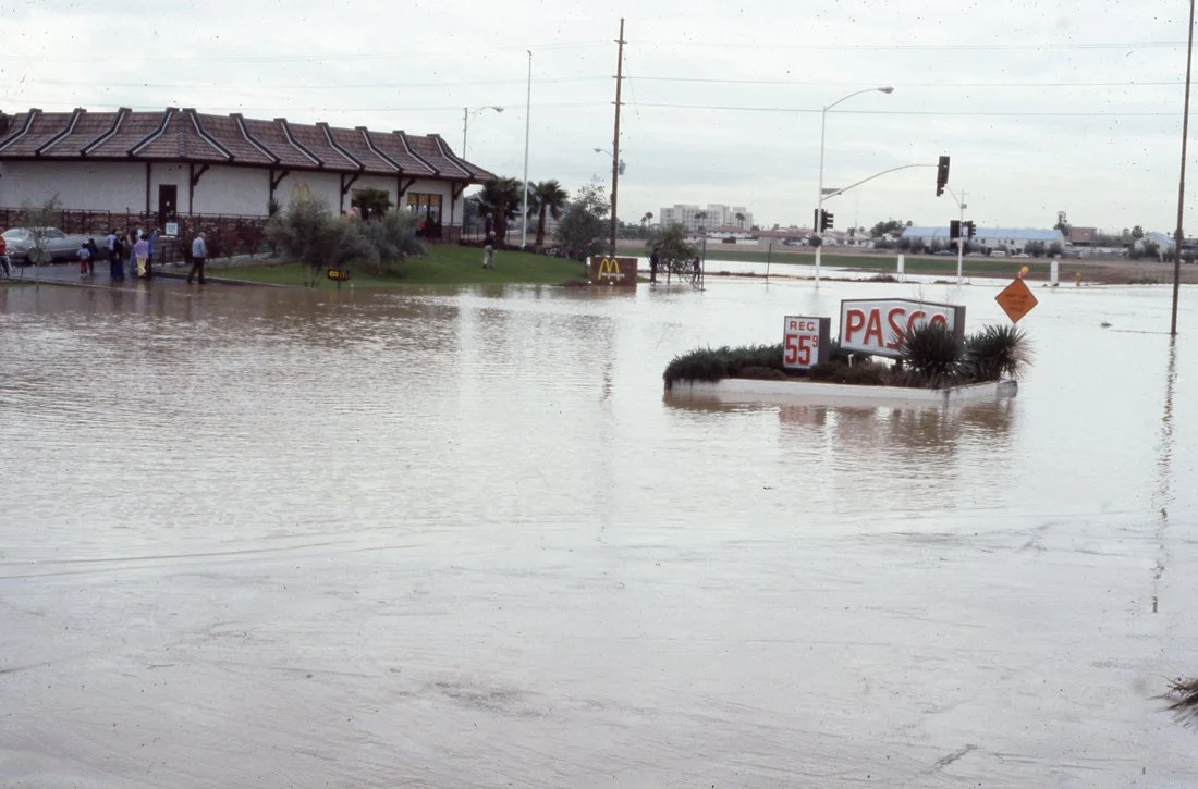 Flooded street with water covering the road and surrounding areas, partially submerging traffic signs and a roundabout with some bushes.