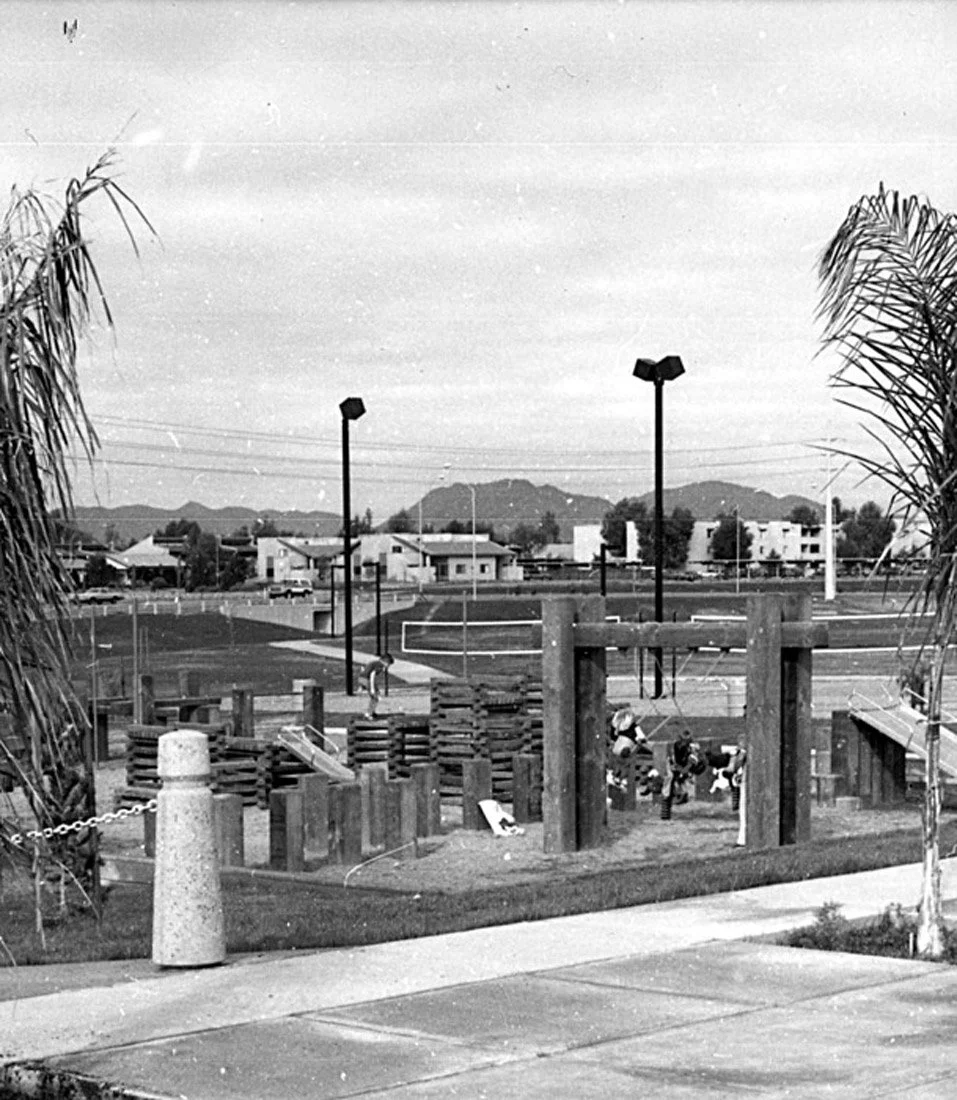 Black and white photo of a playground with swings, wooden climbing structures, and a sandy area, surrounded by trees and located near a residential area with houses and a sports field in the background.