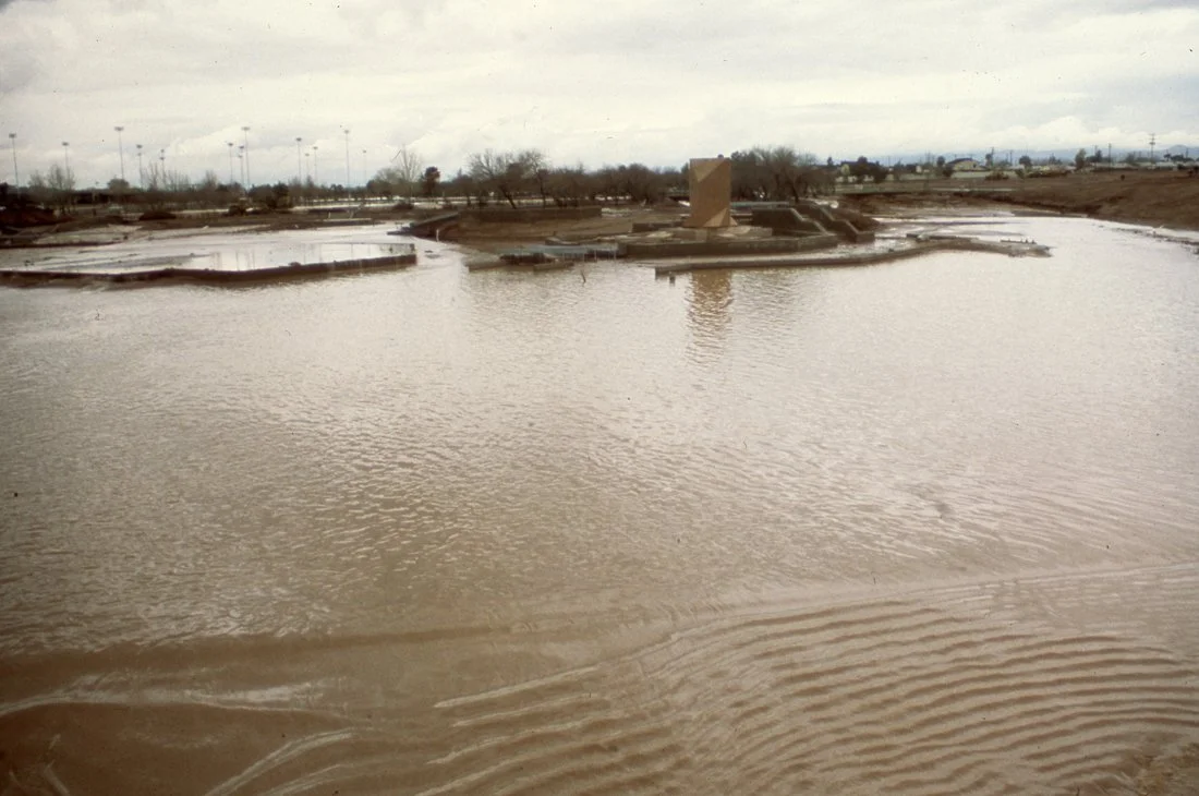 A construction site submerged in floodwaters with muddy water covering the ground and partially flooding structures. In the background, there are trees and light poles under an overcast sky.