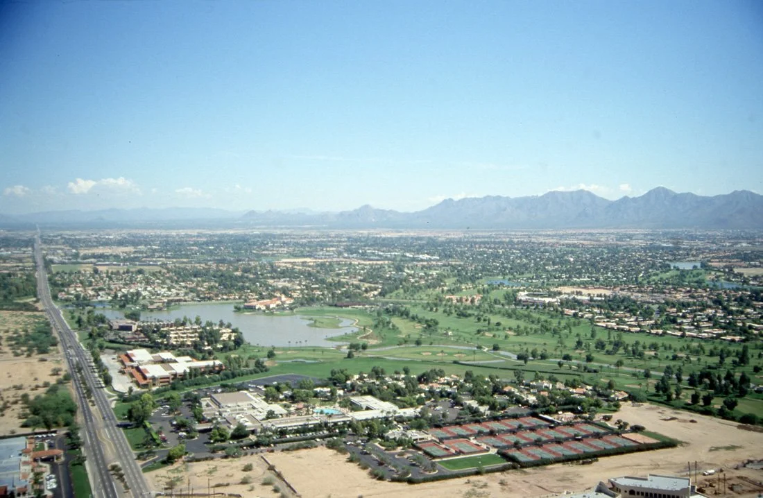 Aerial view of a city with a golf course, bodies of water, and mountains in the background.