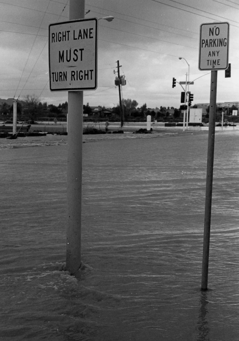 Street signs above floodwaters, one indicating right lane must turn right, and the other indicating no parking at any time.