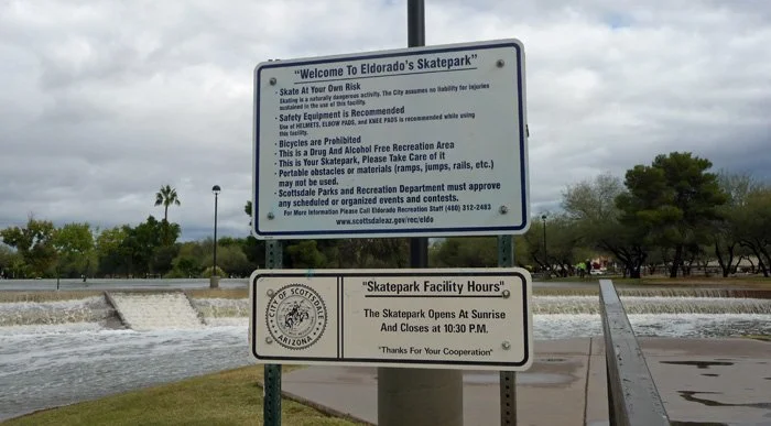 Sign at Eldorado Skatepark with rules and hours, near a small waterfall, trees, and cloudy sky.