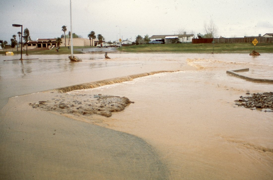 Flooded street with water covering the road, submerged trees, and damaged infrastructure.