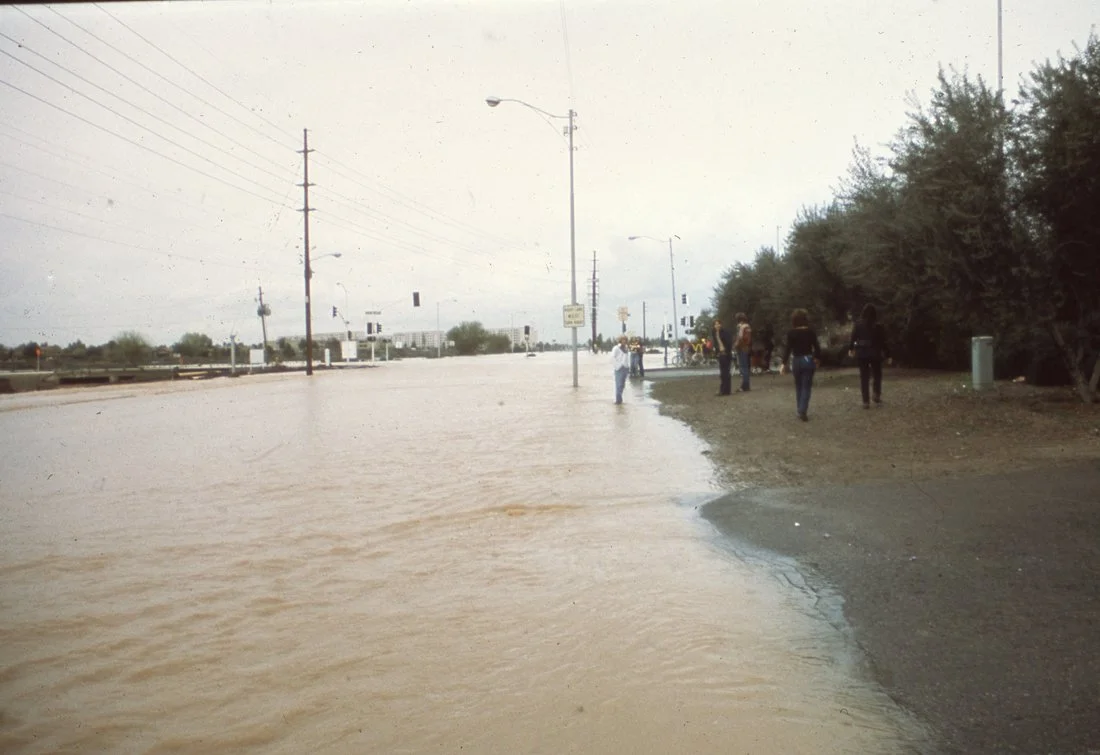 Flooded street with muddy water, pedestrians on the sidewalk, and trees on the right side.
