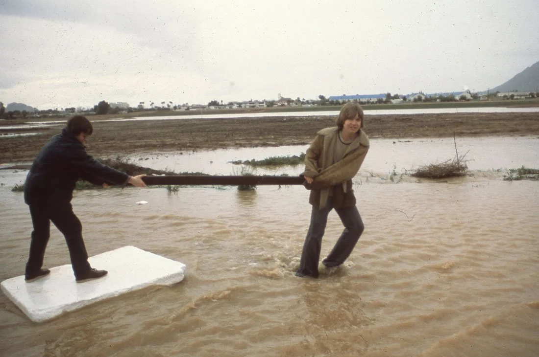 Two young boys play tug-of-war with a long stick in a flooded area during overcast weather.