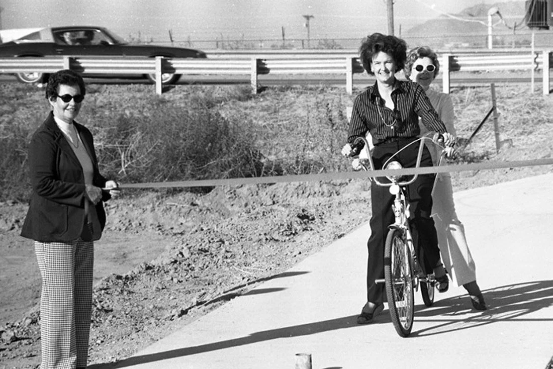 A woman riding a bicycle through a ribbon-cutting ceremony with two other women standing nearby, one holding the ribbon.