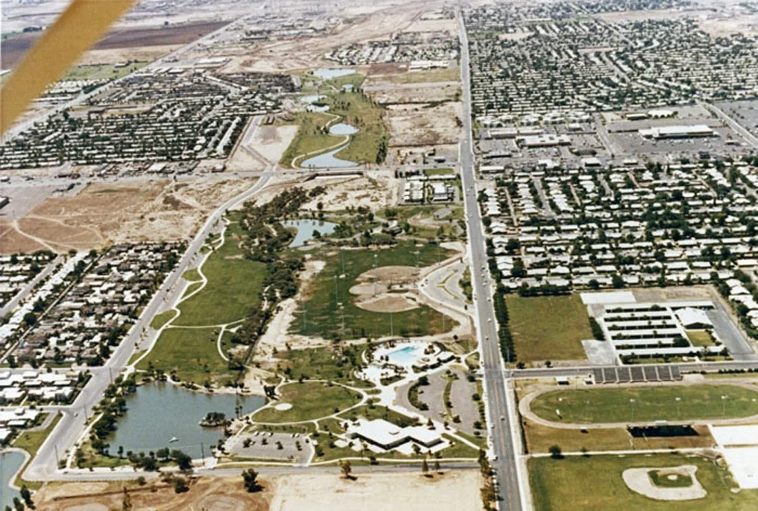 Aerial view of a city park featuring lakes, walking paths, and green spaces, with surrounding residential and commercial areas.