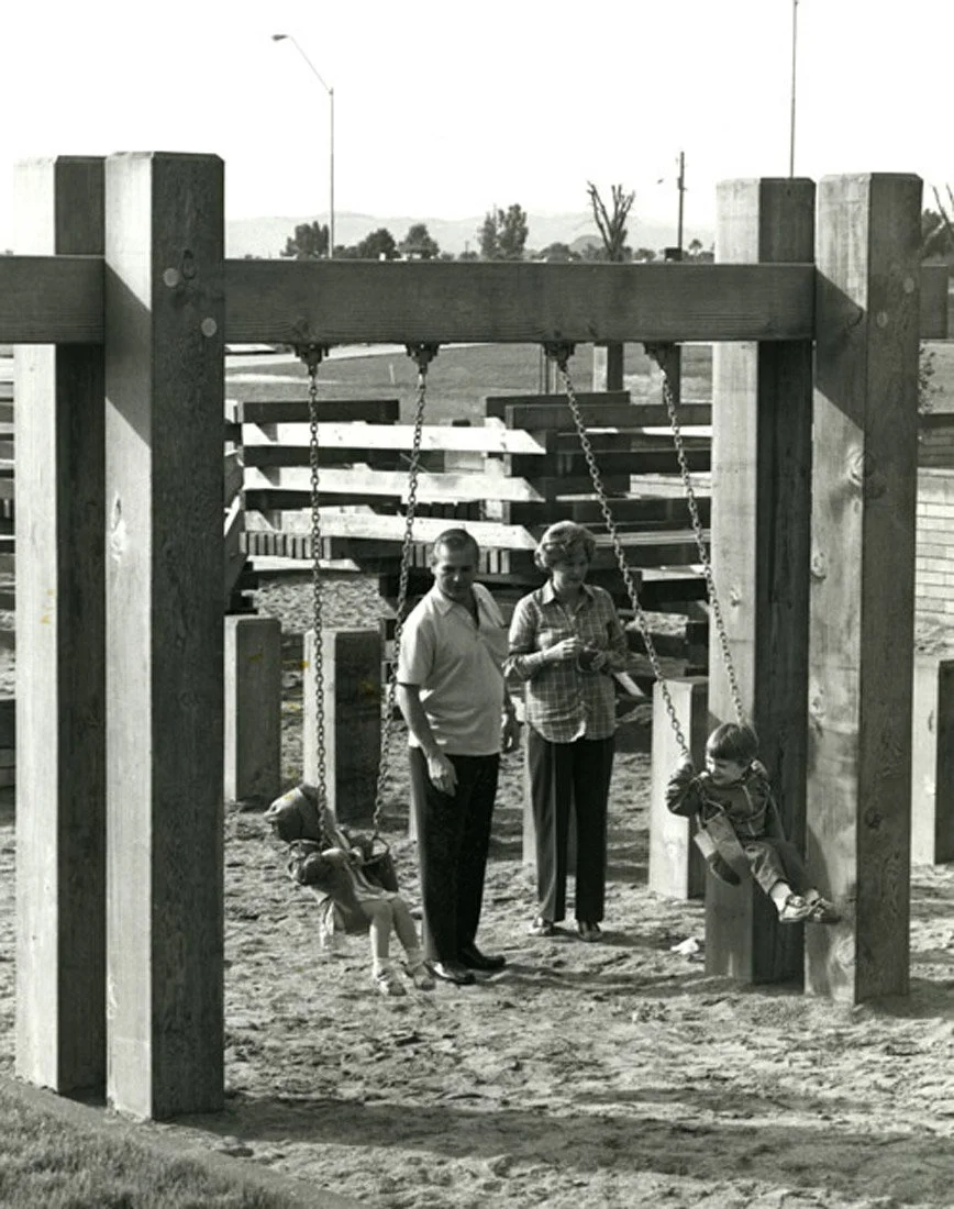 A black and white photograph of a park playground featuring a swing set with two swings. A boy is sitting on one swing and a woman is standing next to the other swing, while a man stands nearby. In the background, there are trees, hills and utility p