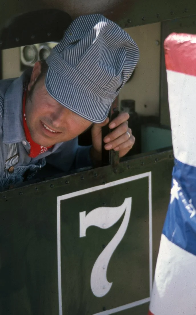 A man wearing a striped cap and denim apron, talking on a phone while leaning out of a vintage green vehicle with the number 7 on it. Part of a flag with red, white, and blue colors is visible on the right.