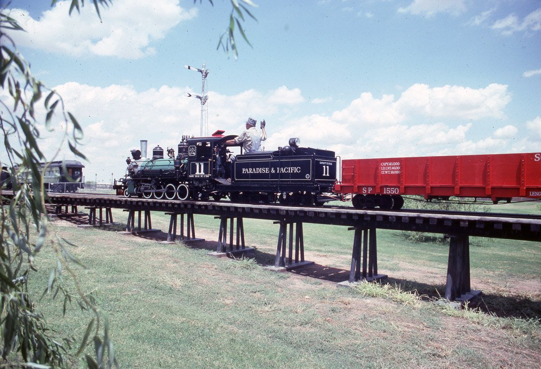 A vintage black steam locomotive with the words "Paradise & Pacific" on its side, pulling a red train car across an elevated track in a grassy area with a blue sky and clouds above.