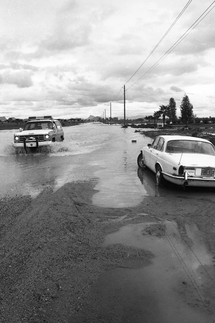 Two cars stuck in a flooded area with water covering the road, under a cloudy sky with power lines overhead.