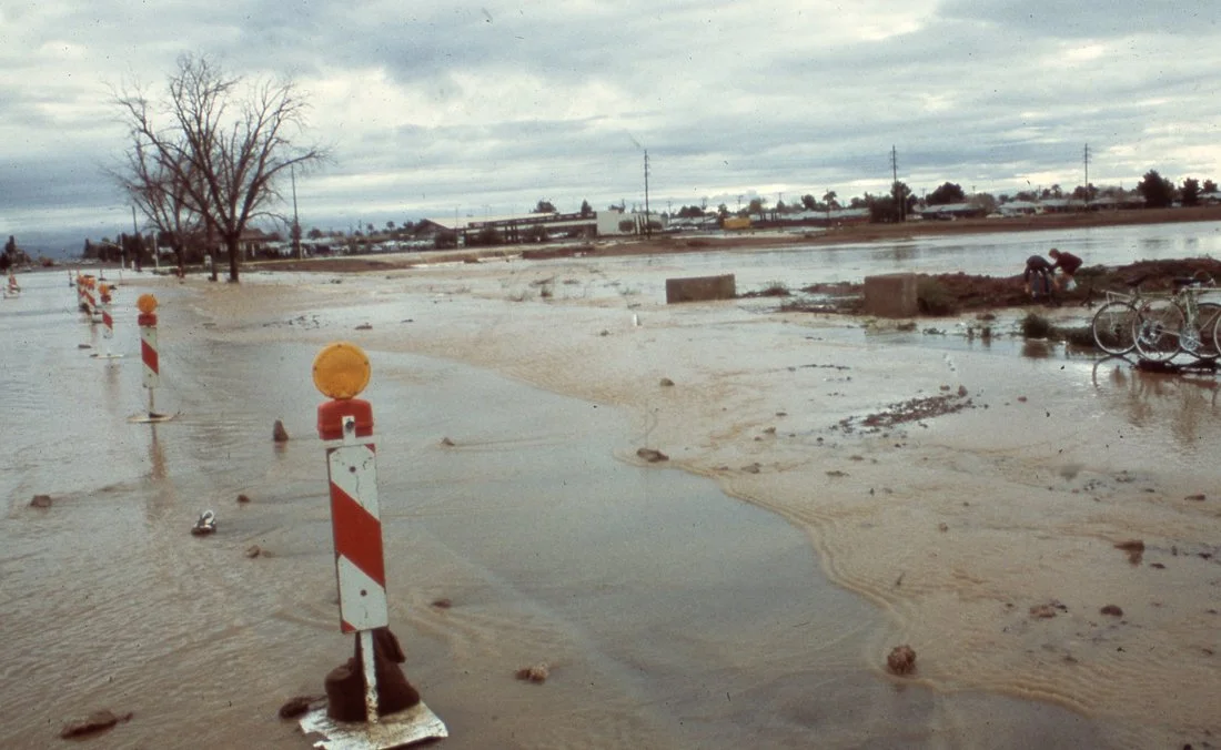 Flooded road with traffic barriers, bicycles, and a person working near the water on an overcast day.
