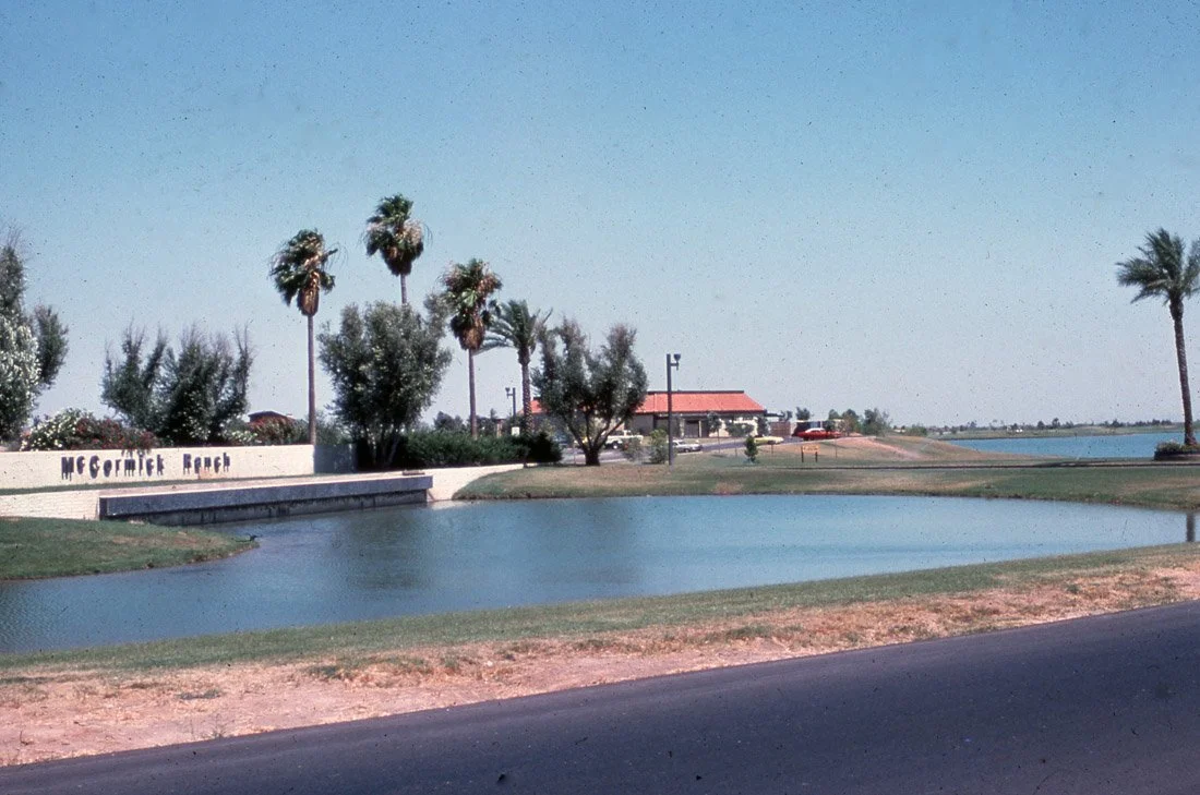 A small lake or pond beside McCormick Ranch in a suburban area with palm trees, a building with a red roof, and a parking lot in the background.
