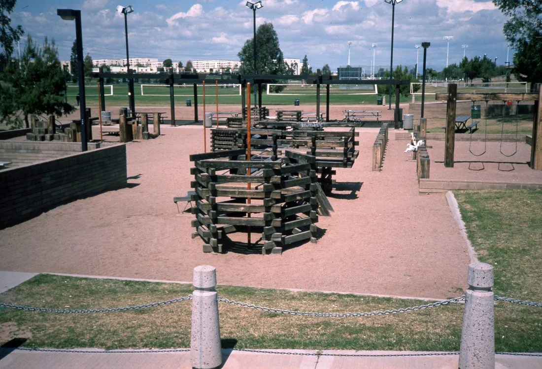 Empty playground with sand surface, wooden structures, swings, and benches on a sunny day.
