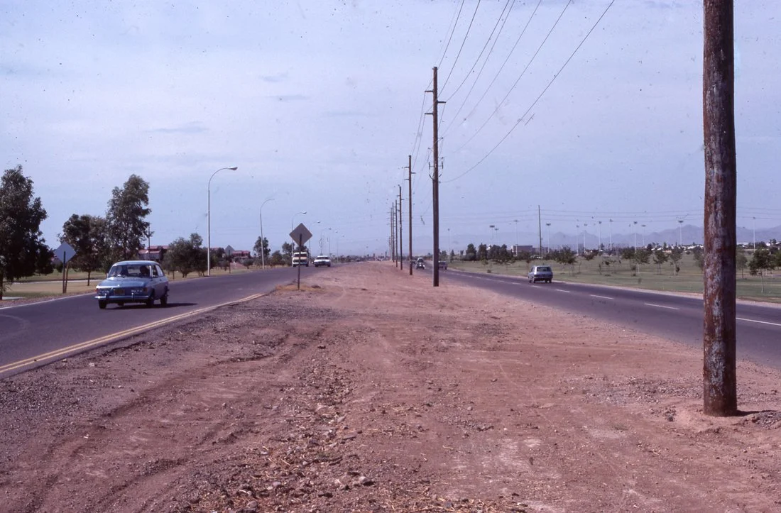 A rural road with cars driving in both directions, utility poles running alongside, and a grassy area with trees on the right. The sky is cloudy.