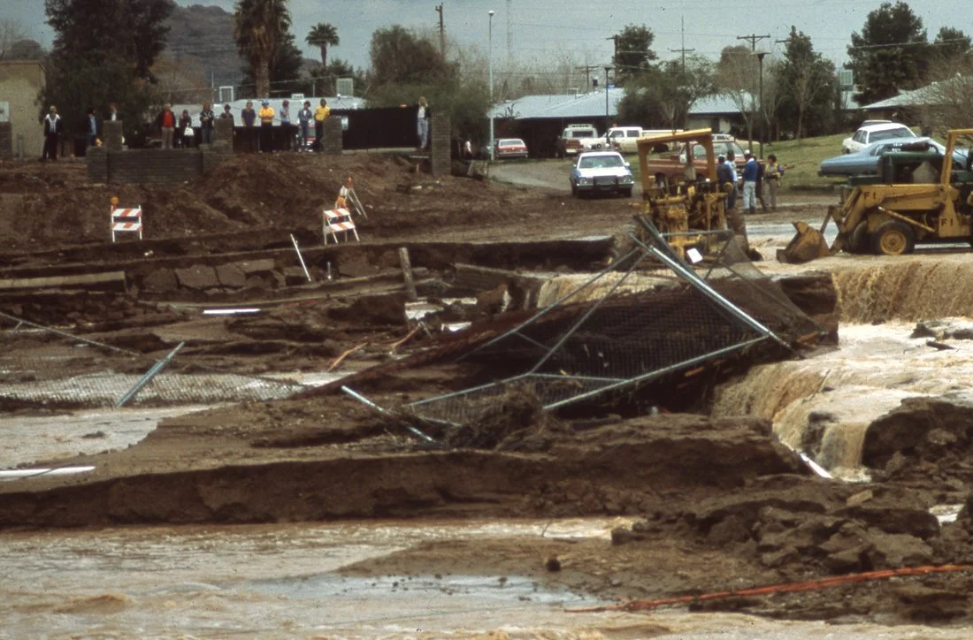 Scene of a large sinkhole on a street, with damaged fencing and construction equipment, surrounded by onlookers, vehicles, and trees in the background.
