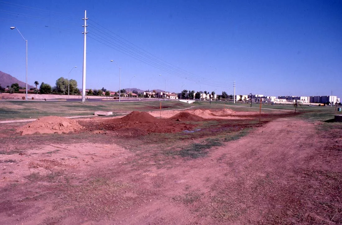 Construction site on a dirt road with small mounds of dirt, power lines, streetlights, and residential buildings in the background under a clear blue sky.