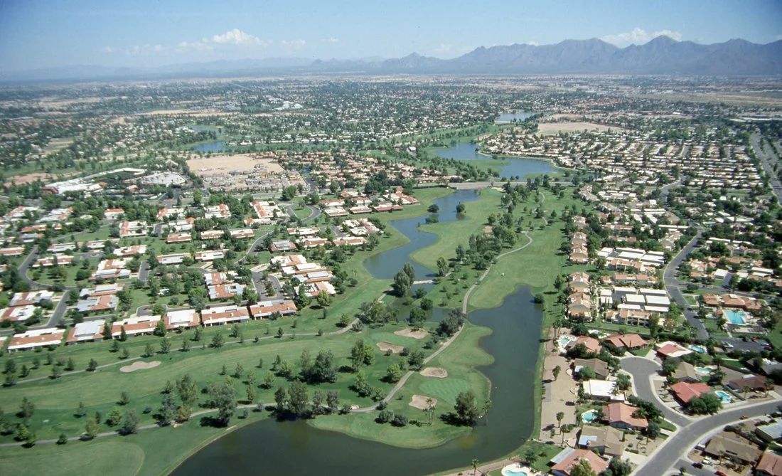 Aerial view of a golf course with houses and water features in a suburban neighborhood, mountains in the background under a blue sky.