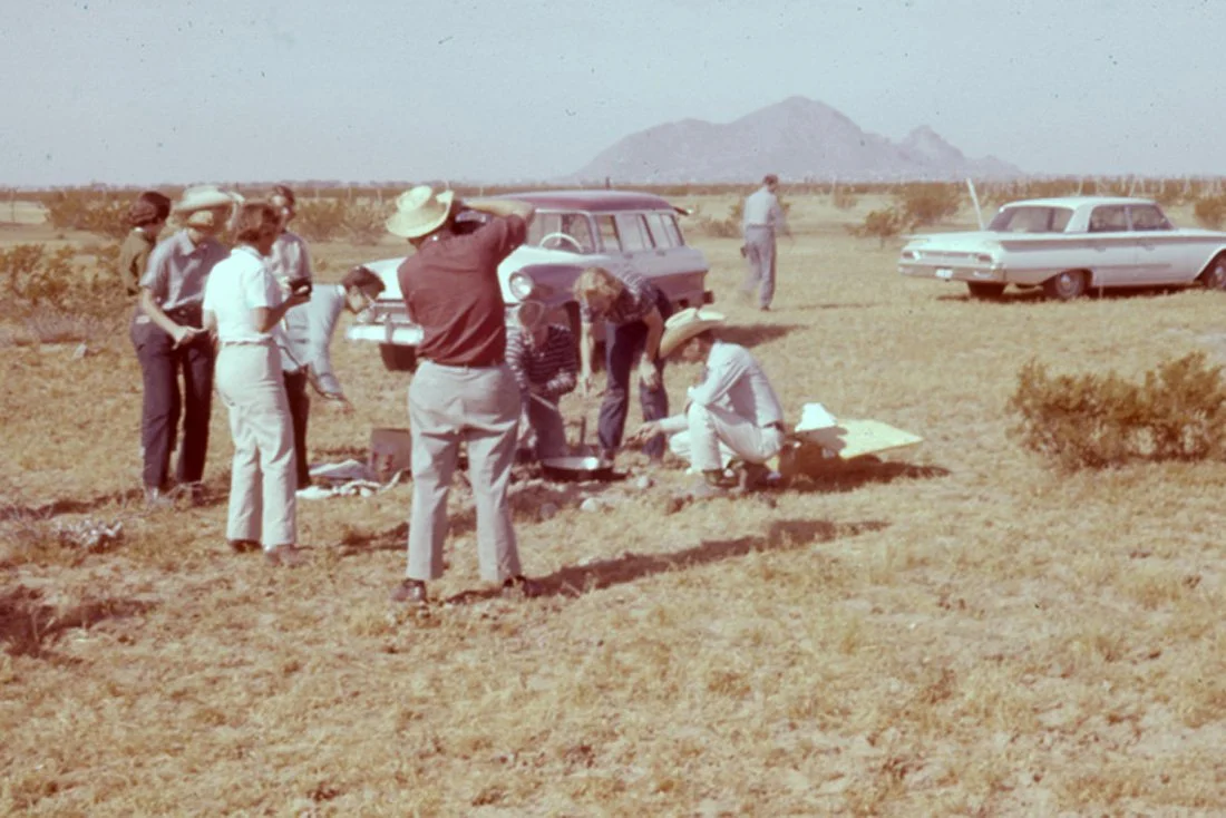 A group of people, some in hats, gather around digging in a dry, open field with vintage cars parked nearby, and mountains in the background.