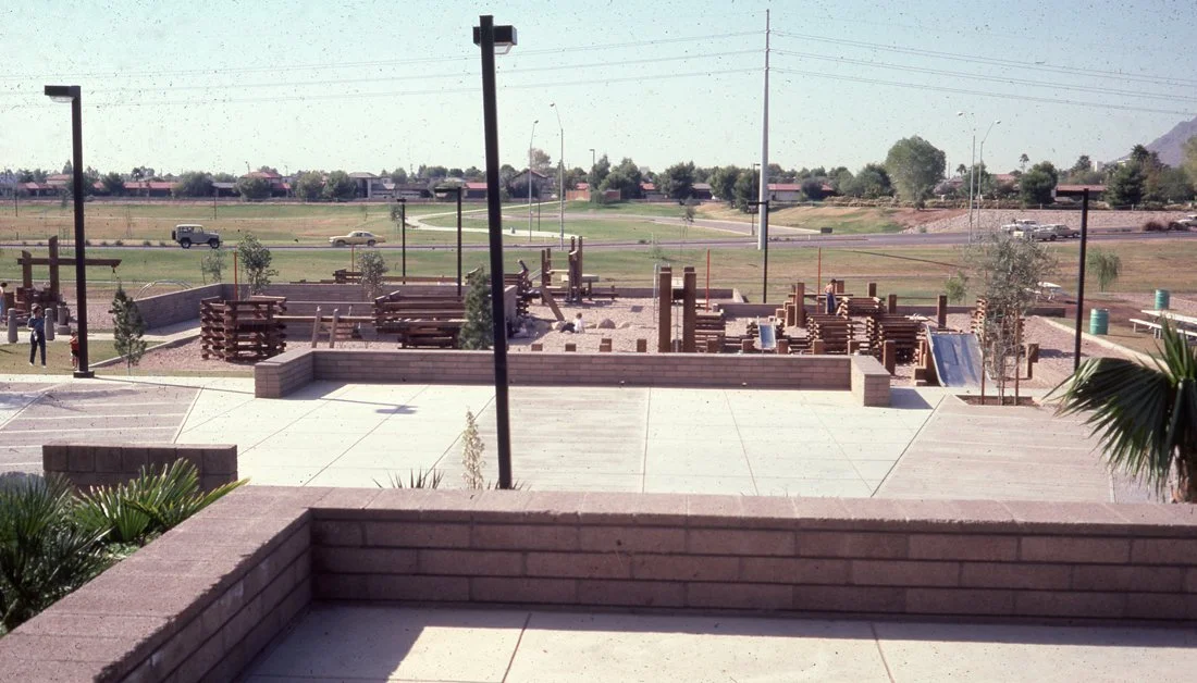 Construction site with stacks of wooden planks and workers, viewed from a paved area with planters and small trees, near a highway with cars passing by in a desert-like area.