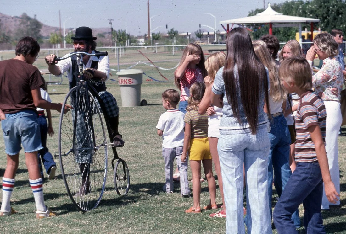 A man dressed as a circus clown riding a tall unicycle while performing for children at an outdoor event, with a group of kids and adults watching.