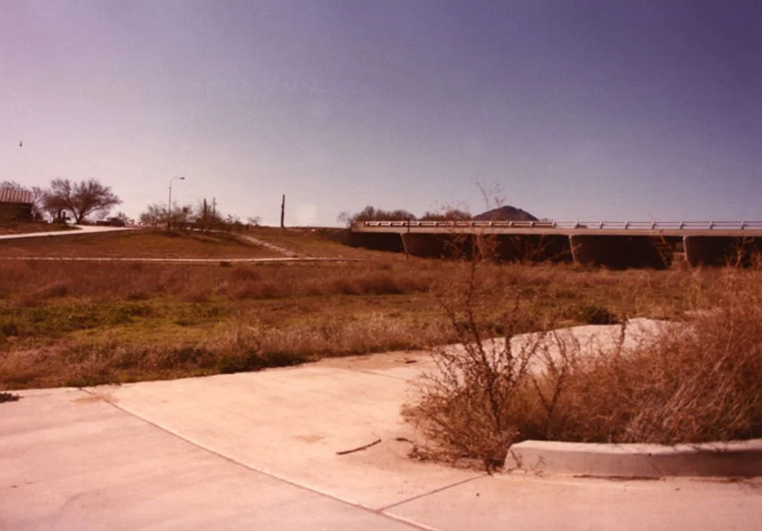 A dry, grassy landscape with a small concrete sidewalk in the foreground, sparse bushes, an overpass for a highway, a few trees, and a clear sky.