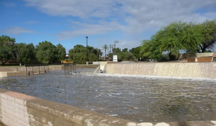 A river or canal with water overflowing a low stone wall, with trees and a partly cloudy blue sky in the background.