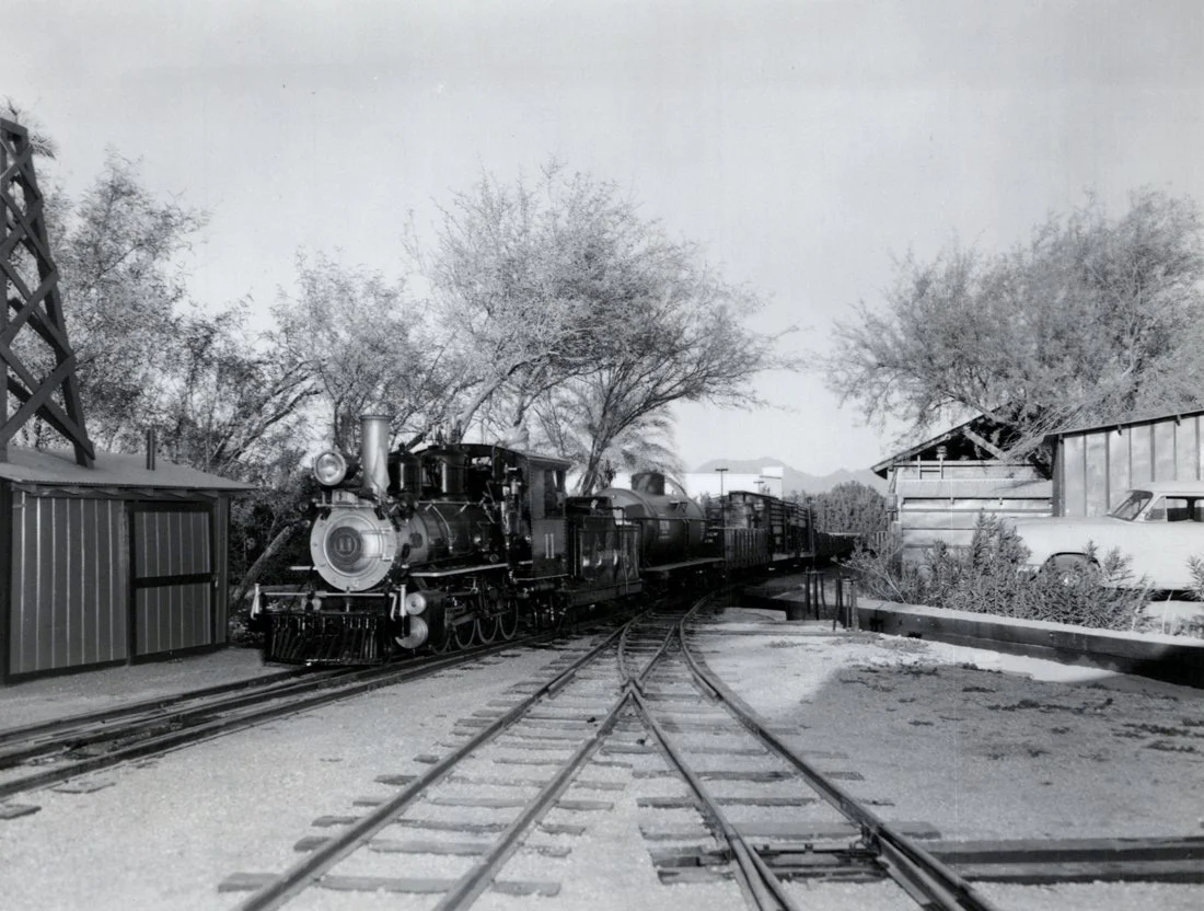 Black and white photo of a vintage steam locomotive train on railway tracks, with trees and a building in the background.