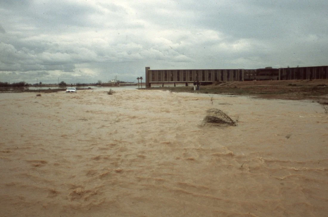 Flooded area with muddy water, a few tufts of grass, a building in the background, and cloudy sky.