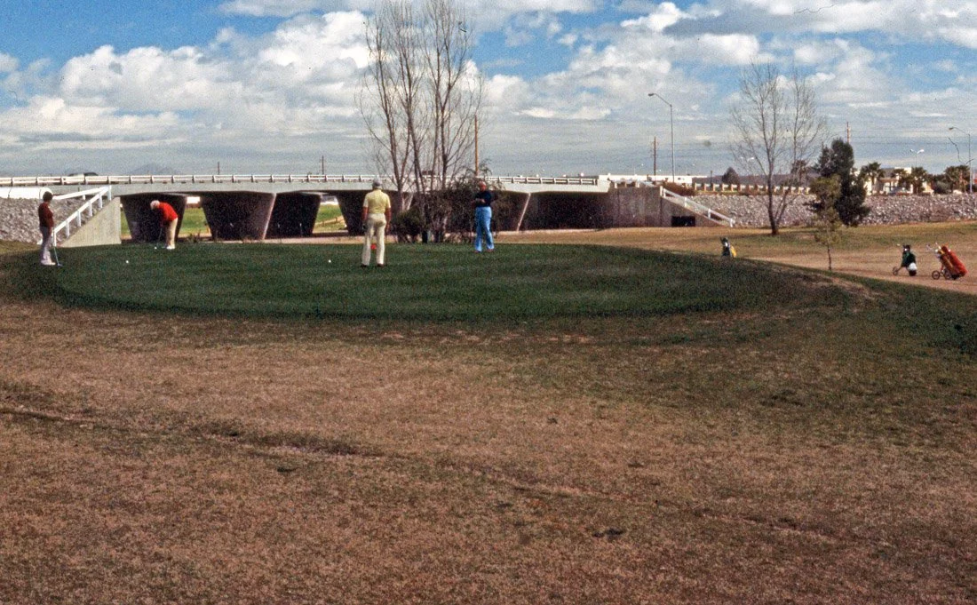 Golfers practicing putting on a green near a bridge on a partly cloudy day.