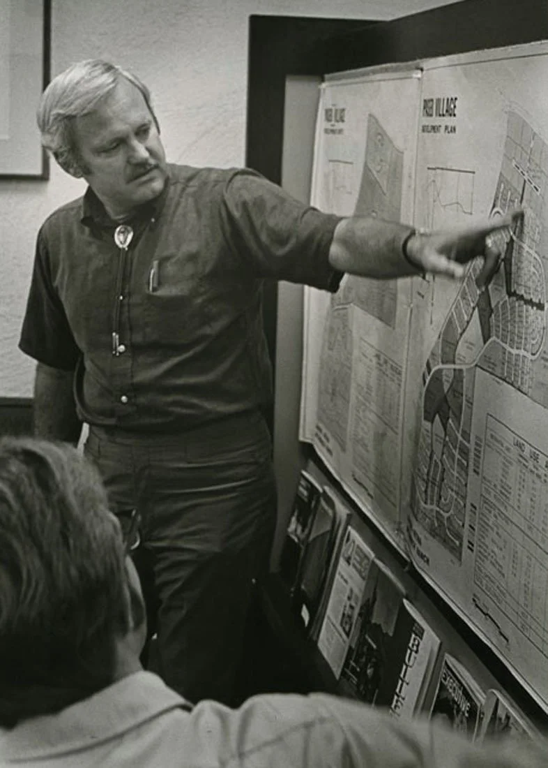 A man with light-colored hair wearing a short-sleeved button-up shirt and a bolo tie is pointing at a large detailed map on a display board, with two seated men observing, in a black and white photo.