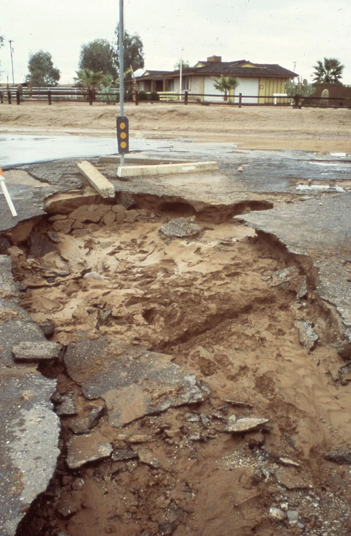 A large pothole in the middle of a damaged road with exposed dirt and broken asphalt. A pole with reflectors and wooden planks are nearby. In the background, there is a house with a yard and trees.