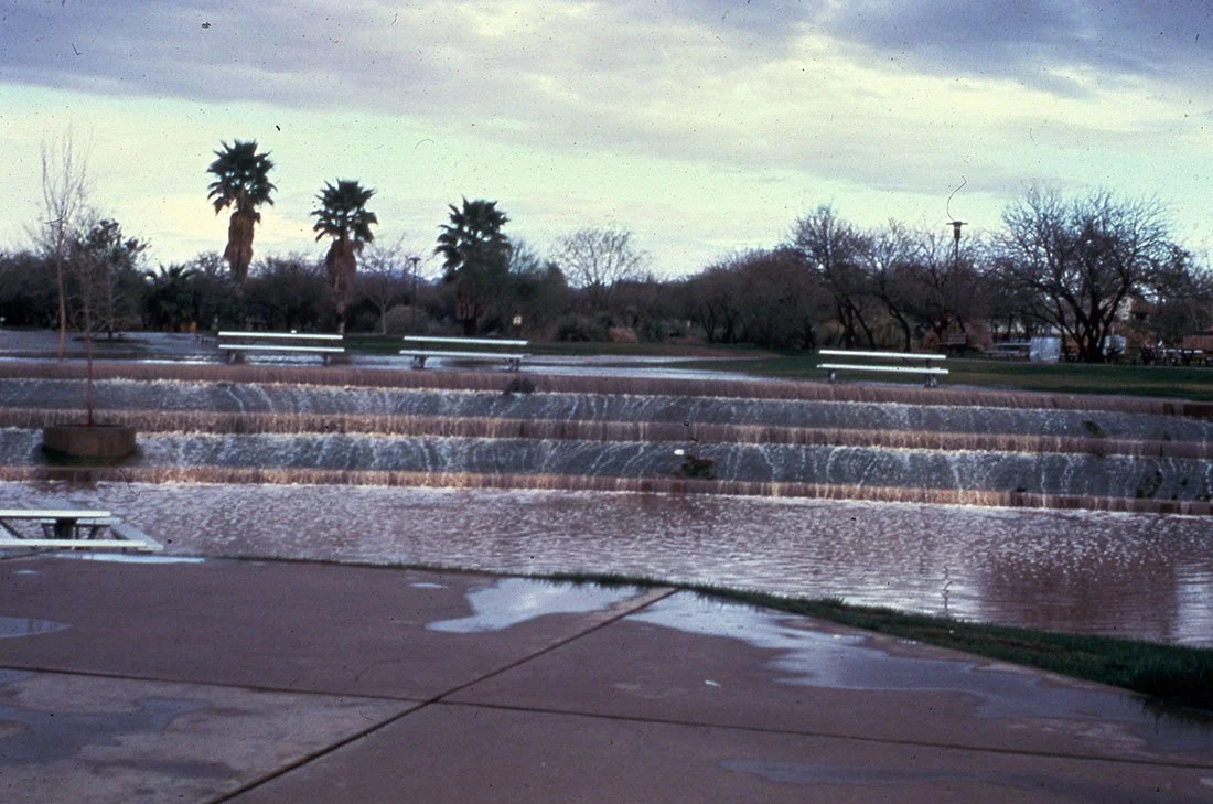 A park with a small waterfall, water flowing over rocks, and benches nearby, with trees and palm trees in the background under a cloudy sky.