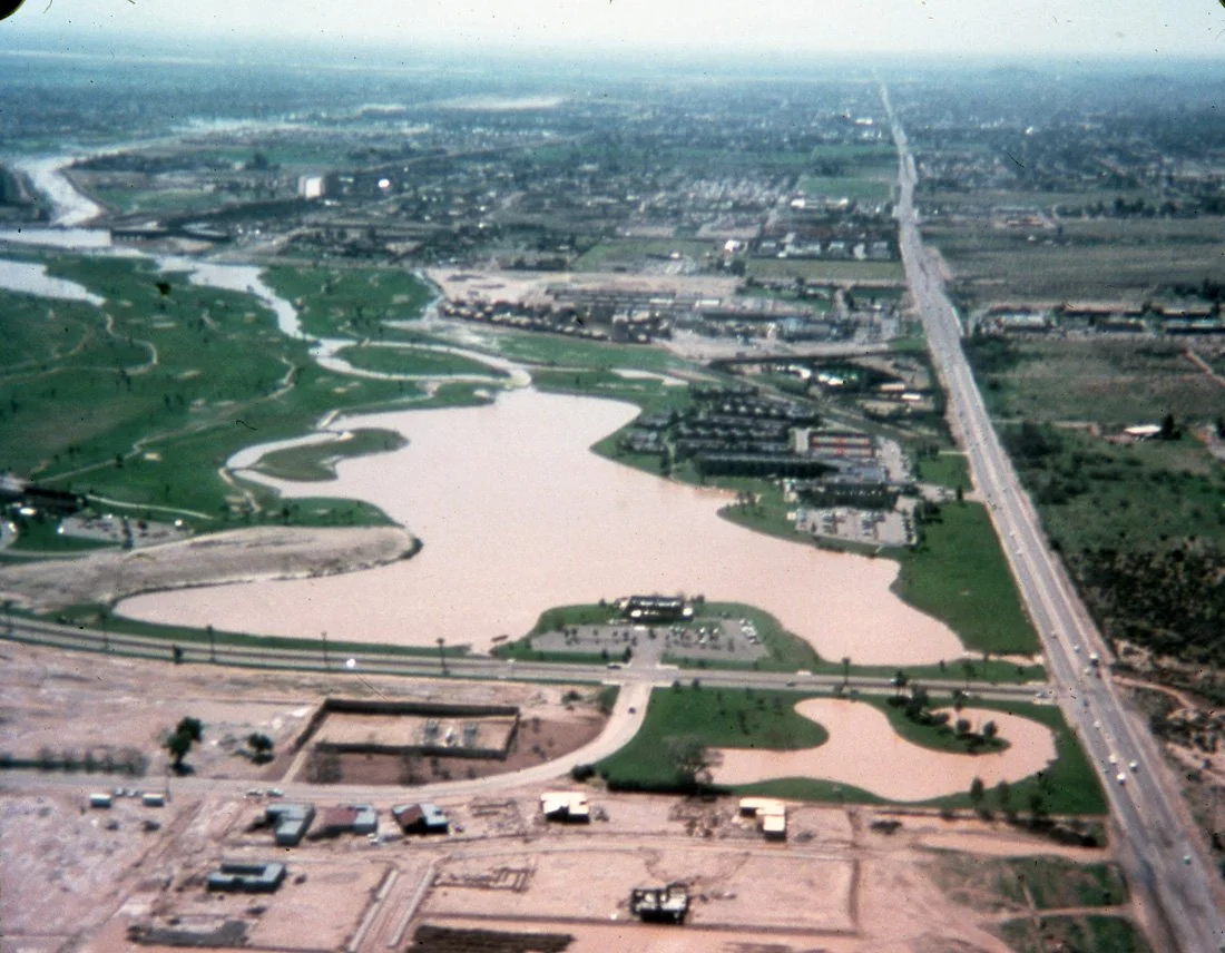 Aerial view of a city park with water features, winding pathways, and surrounding roads and buildings.